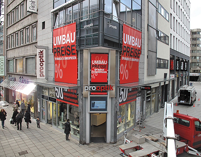 Pro Optik Königstrasse StuttgartPVC Banner. Temporäre Aussenwerbung während der Umbauphase. Zur Montage wurden oben und unten Auluminiumstangen durch Hohlsäume am Banner geschoben. Die seiten wurden mittels Ösen an einem vertikal laufenden Stahlseil verspannt.